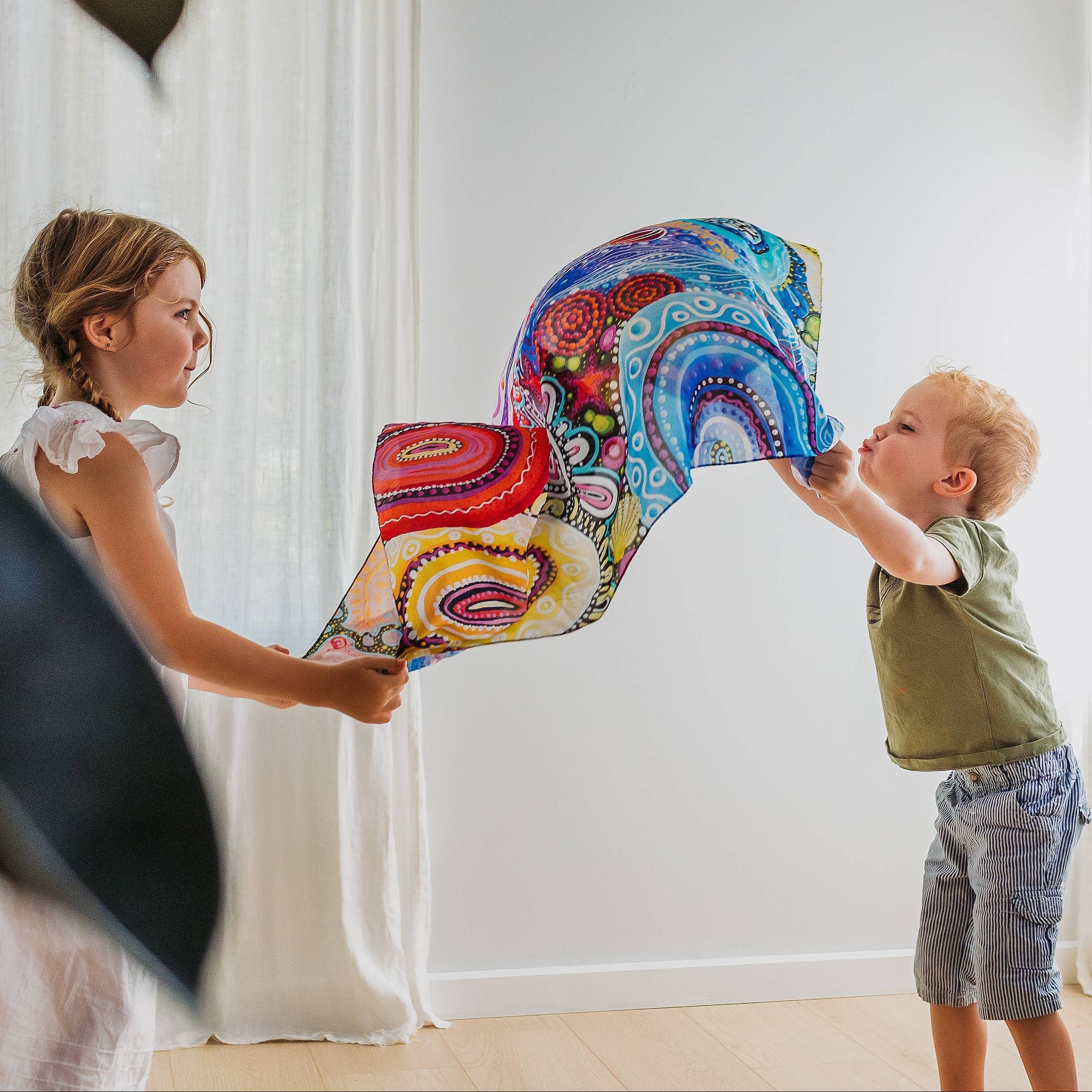 Two children playing with a colorful silk in a room with white curtains.