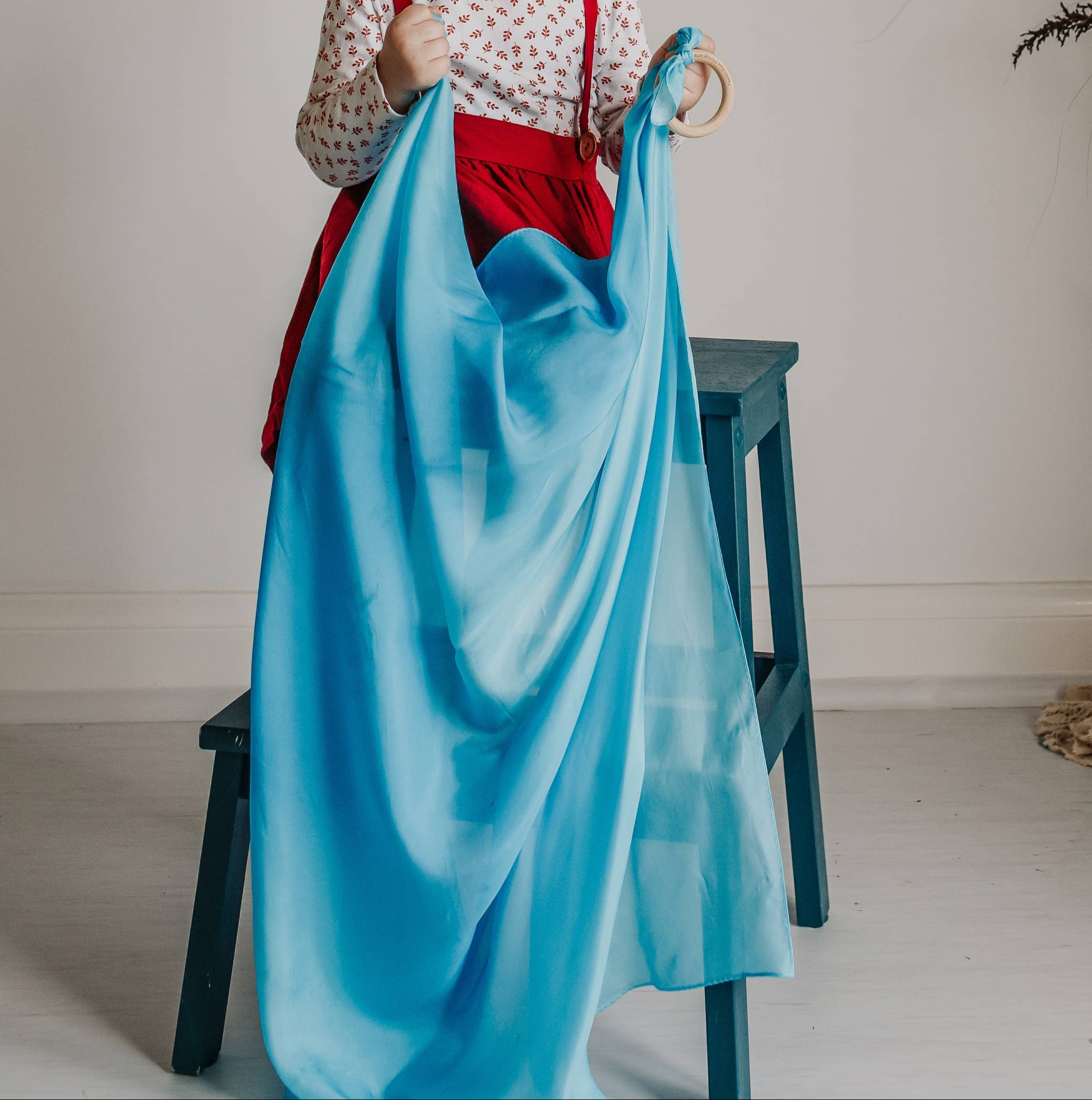 Child holding a blue fabric in a room with a plant and stool.