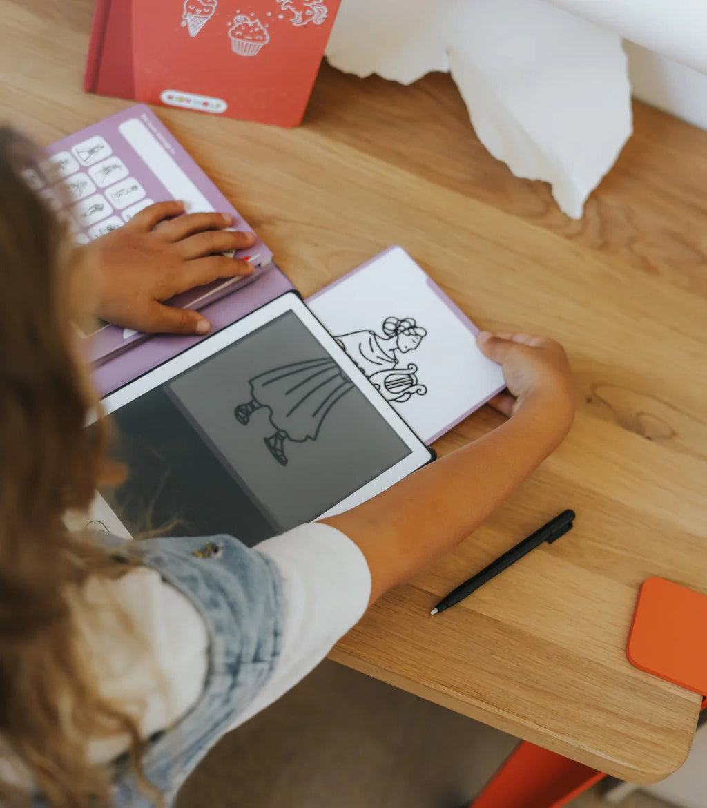 Person using a digital drawing tablet on a wooden table with a pen and paper.