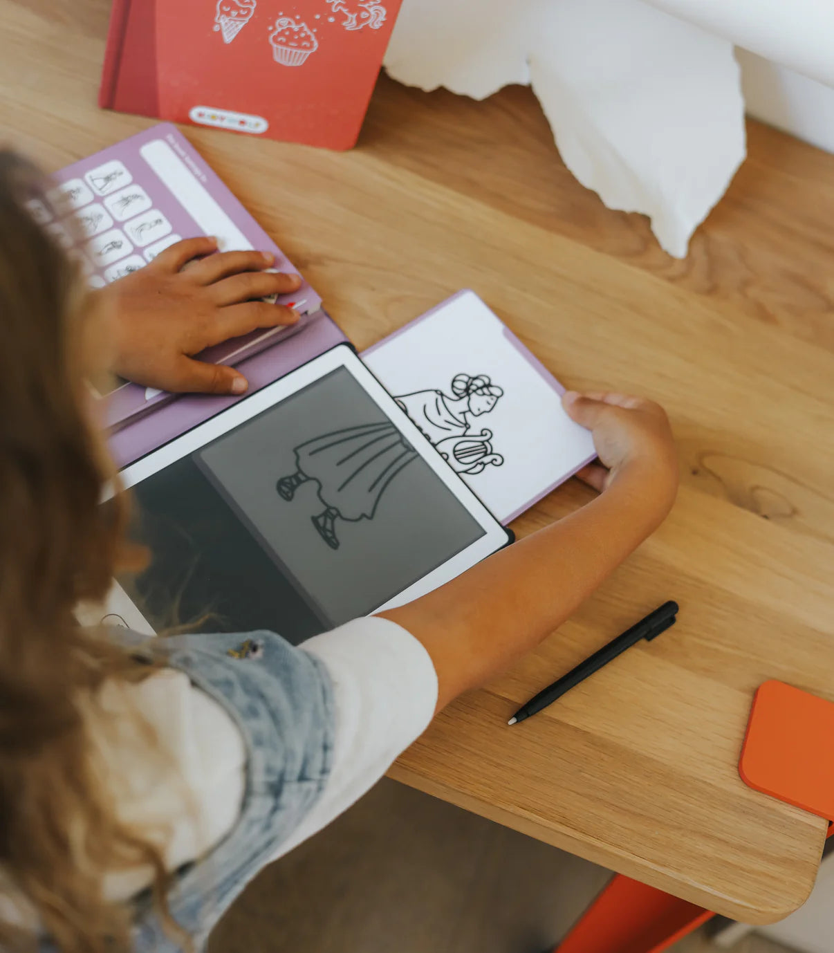 Person using a digital drawing tablet on a wooden table with a pen and paper.