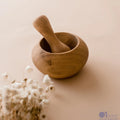 A wooden pestle resting inside a wooden mortar, with a bunch of dried white flowers next to it, all placed on a beige background.