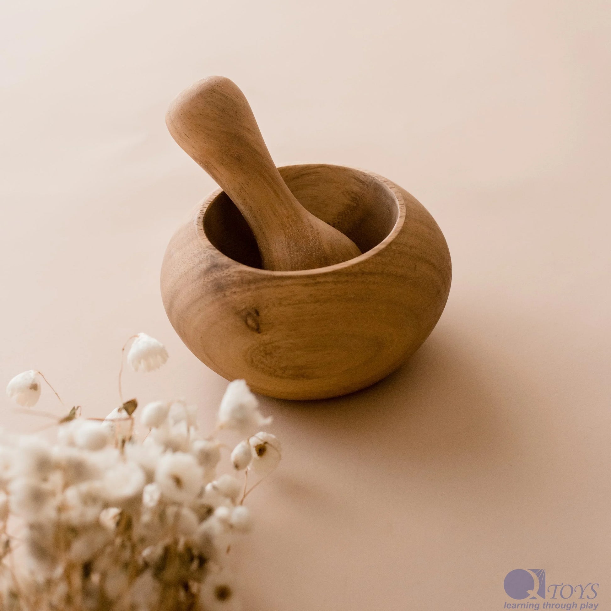 A wooden pestle resting inside a wooden mortar, with a bunch of dried white flowers next to it, all placed on a beige background.