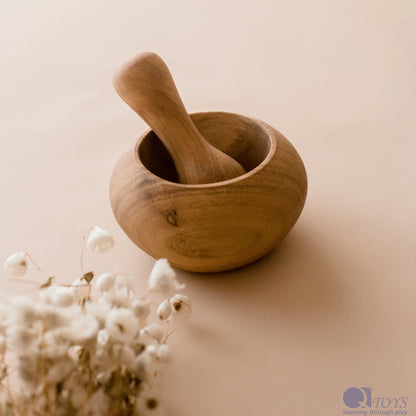A wooden pestle resting inside a wooden mortar, with a bunch of dried white flowers next to it, all placed on a beige background.