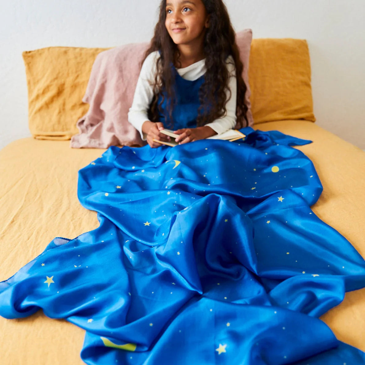 Child sitting on bed with yellow sheets and starry night play silk draped over their legs.