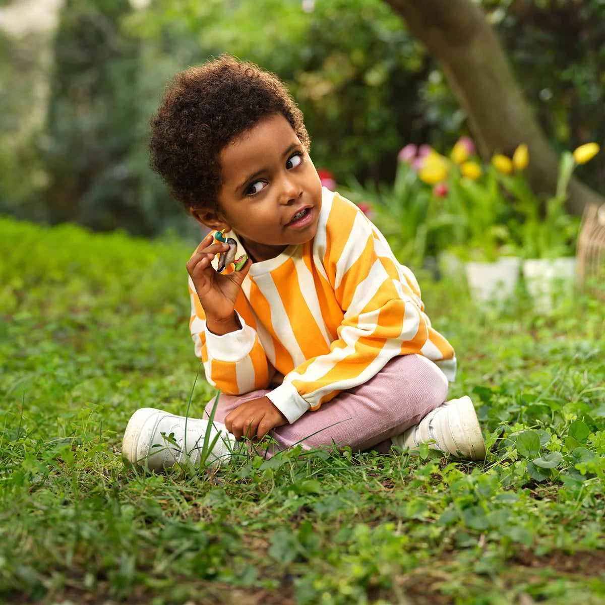 Child sitting on grass holding a toy, surrounded by flowers and greenery