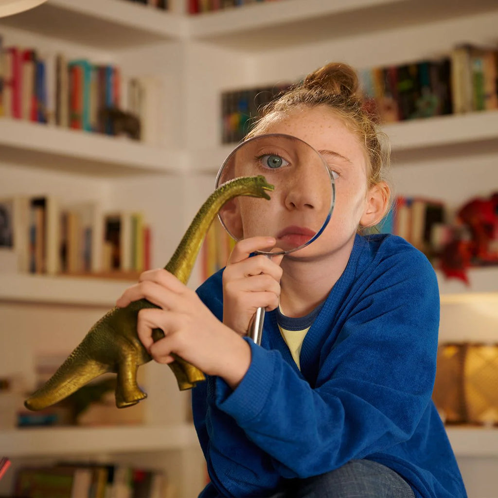 Child holding a dinosaur toy and magnifying glass in a room with bookshelves.