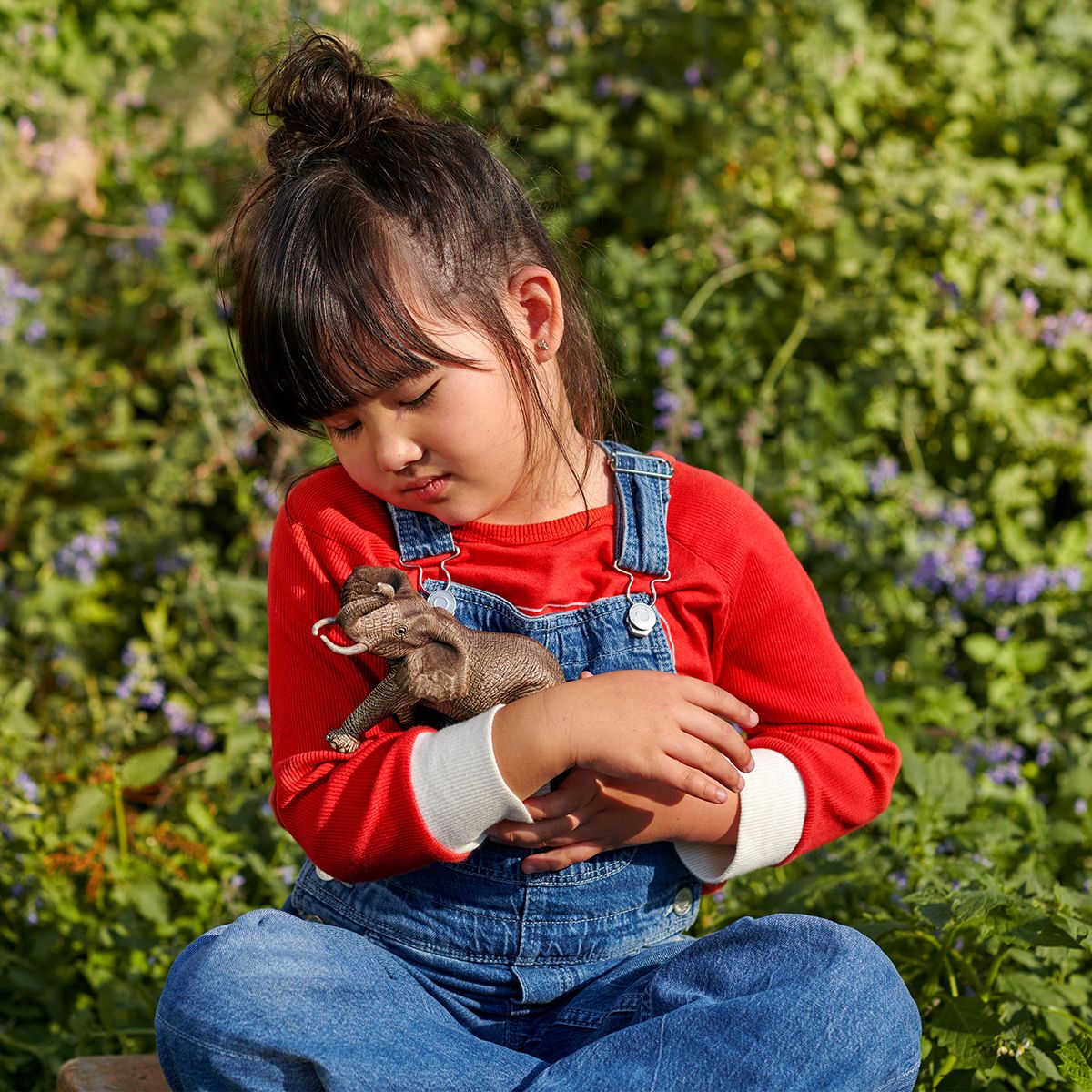 Child holding a small animal in a field with greenery and flowers in the background