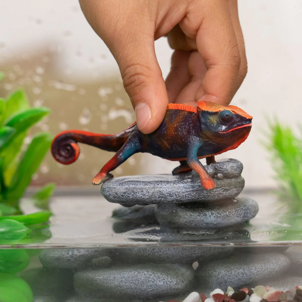 Colorful chameleon figurine on a rock in an aquarium setting with a hand holding it.