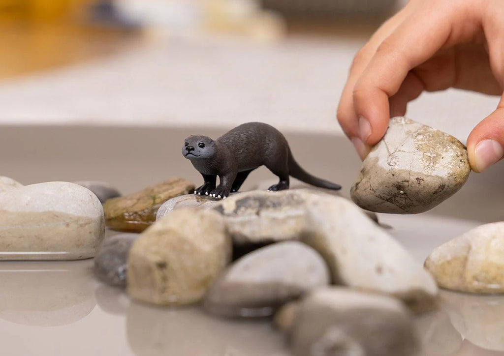 Toy otter on a pile of stones with a hand holding a real stone