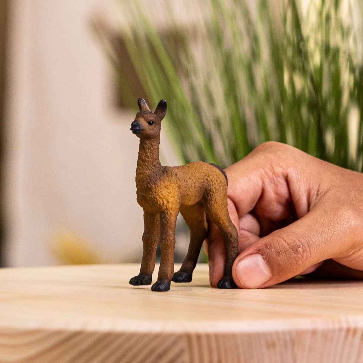 Hand holding a small brown llama figurine on a wooden surface with blurred greenery in the background