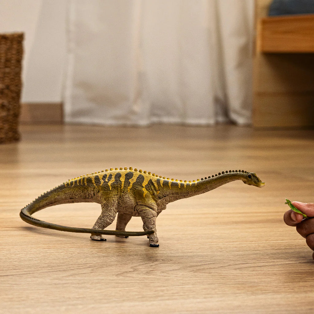 Dinosaur toy figure on a wooden floor with a hand holding a green leaf