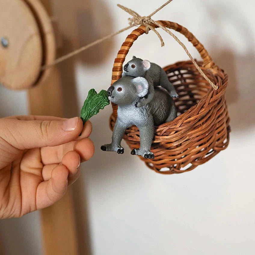 Hand holding a toy koala with a leaf, with another toy koala in a basket.