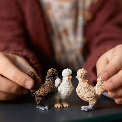 Three small figurines of chickens being held by a person's hands on a dark surface.