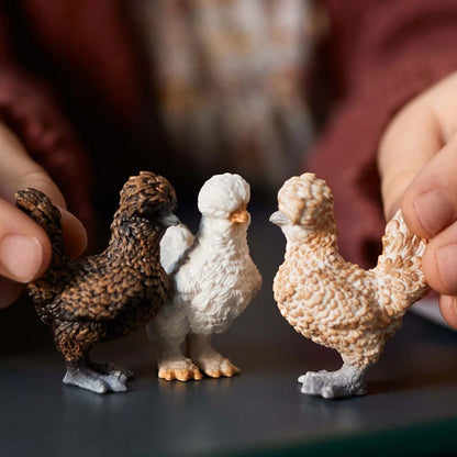 Three figurines of chickens being held by hands on a dark surface.