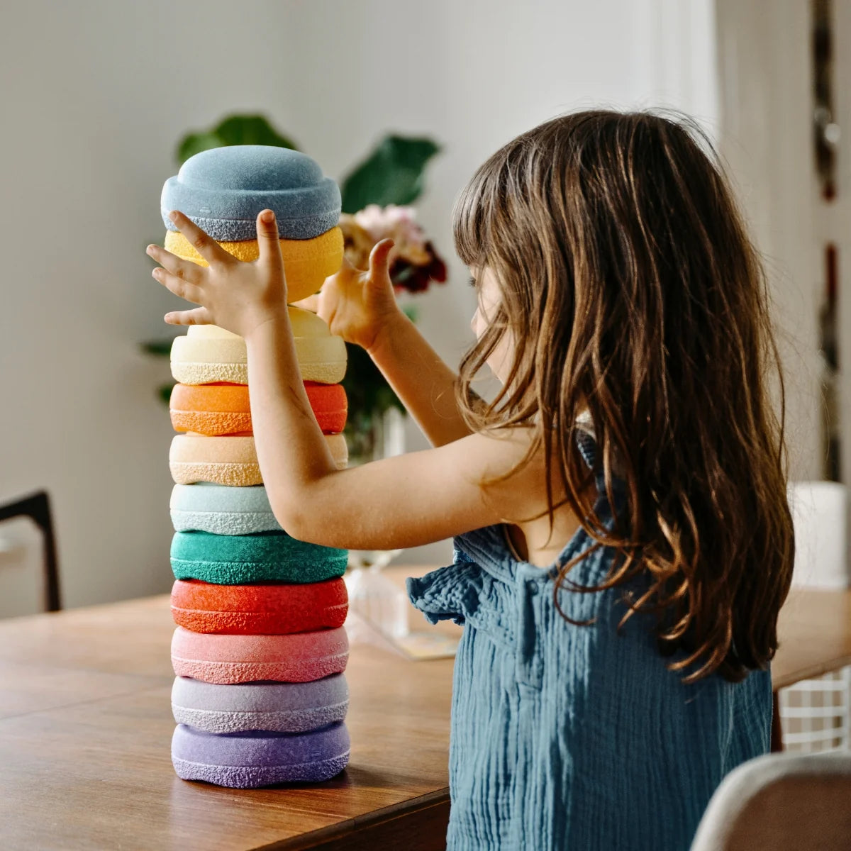 A young child with long brown hair stands at a wooden table, stacking a tall tower of colorful, textured bath towels. The towels are stacked in a rainbow sequence of blue, yellow, cream, orange, pink, red, teal, purple, and light blue.