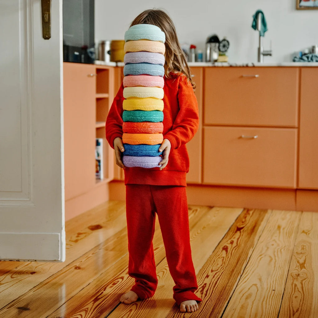 Child in red outfit holding a stack of colorful bowls in a kitchen.