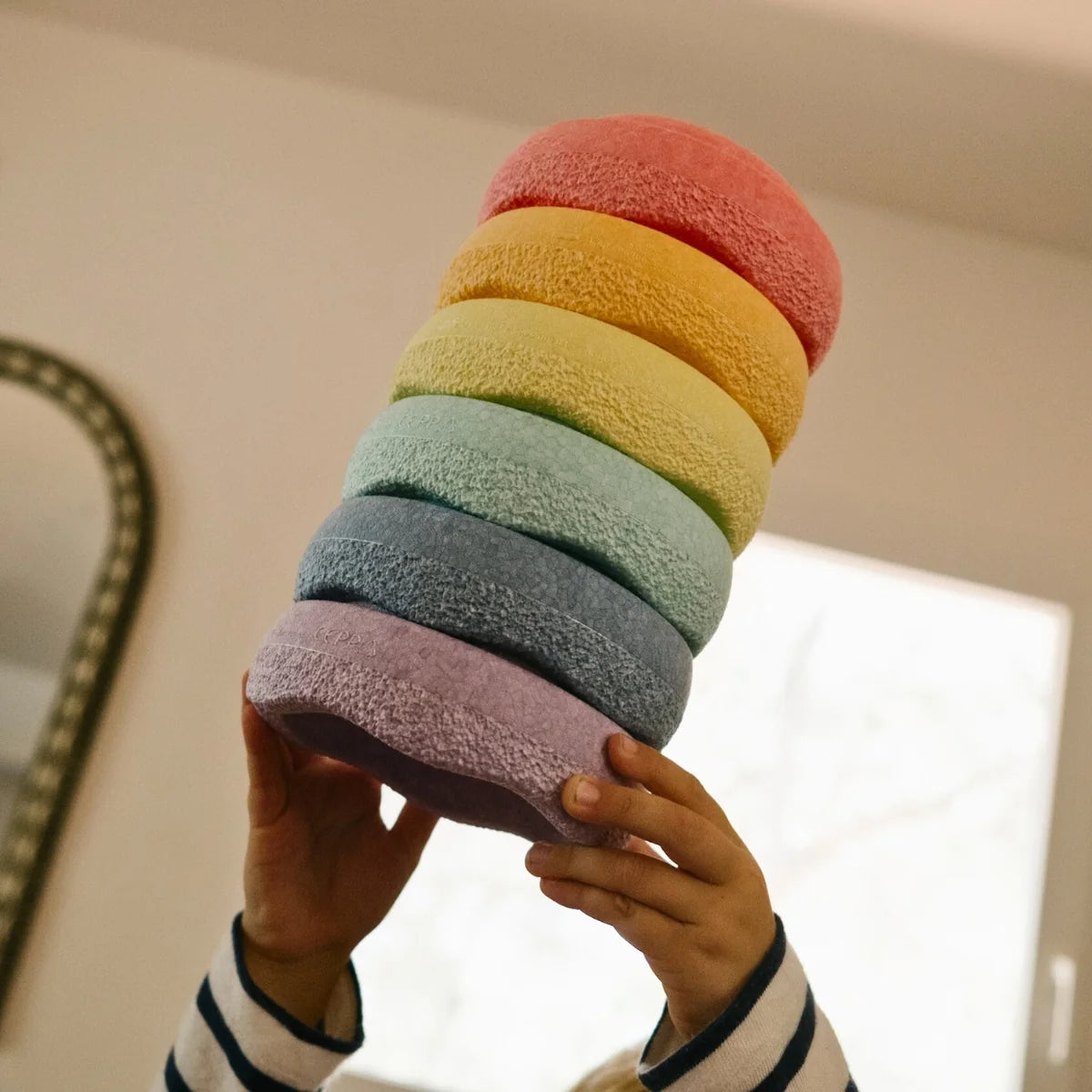 A person holds a stack of mini stepping stones in a rainbow color sequence (pink, orange, yellow, green, blue, purple, and mauve) against a softly lit indoor background.
