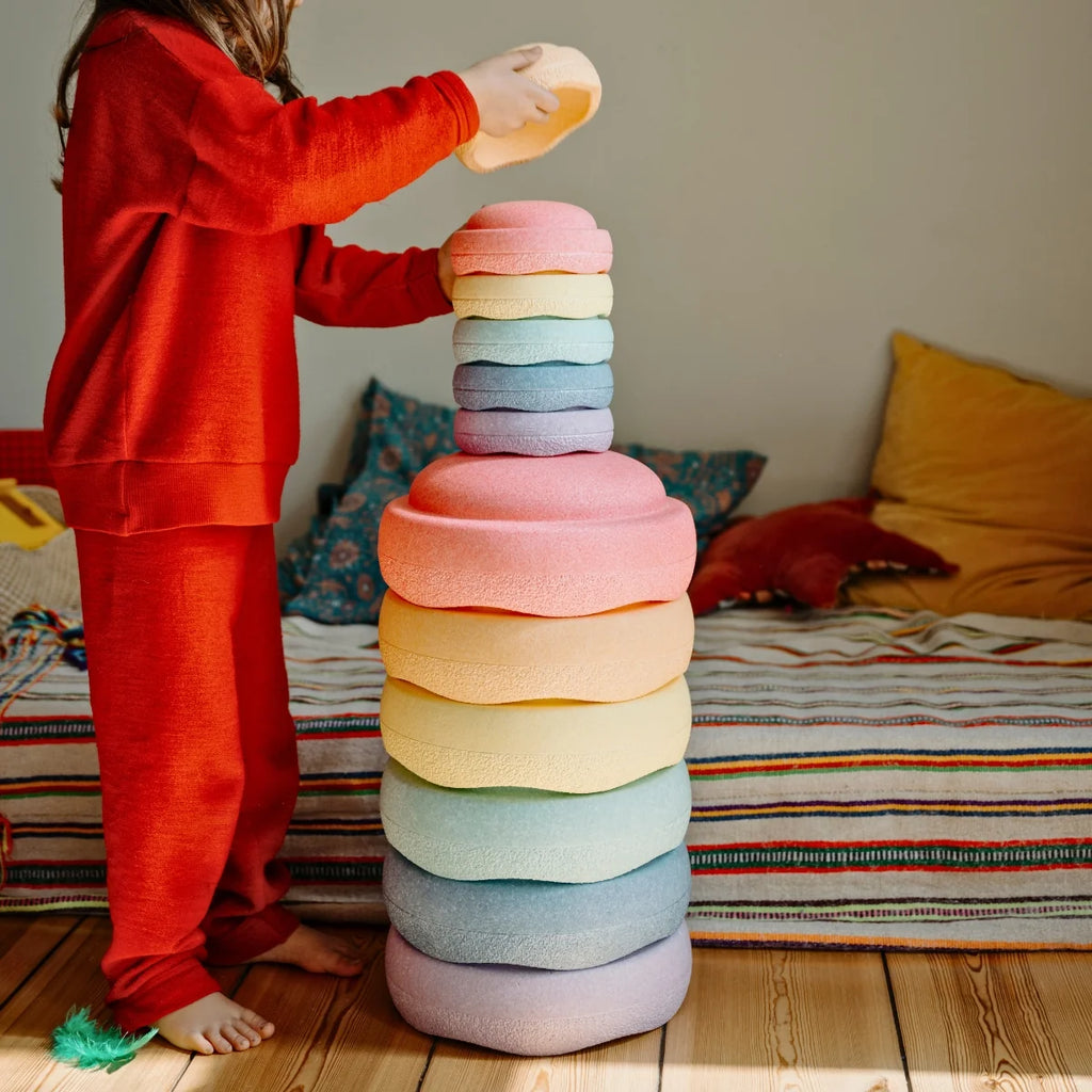 Child in red pajamas stacking colorful stones on a wooden floor.