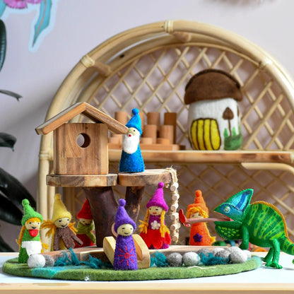 Colorful toy figures of elves and a dinosaur on a wooden base with a wicker chair in the background.