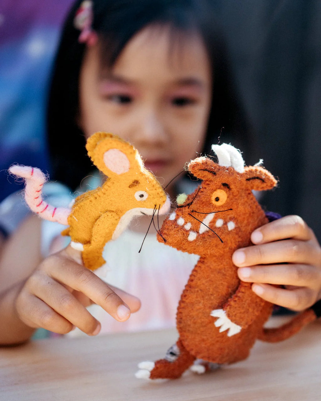 Child holding two felt animals, one orange and one brown, on a wooden surface.