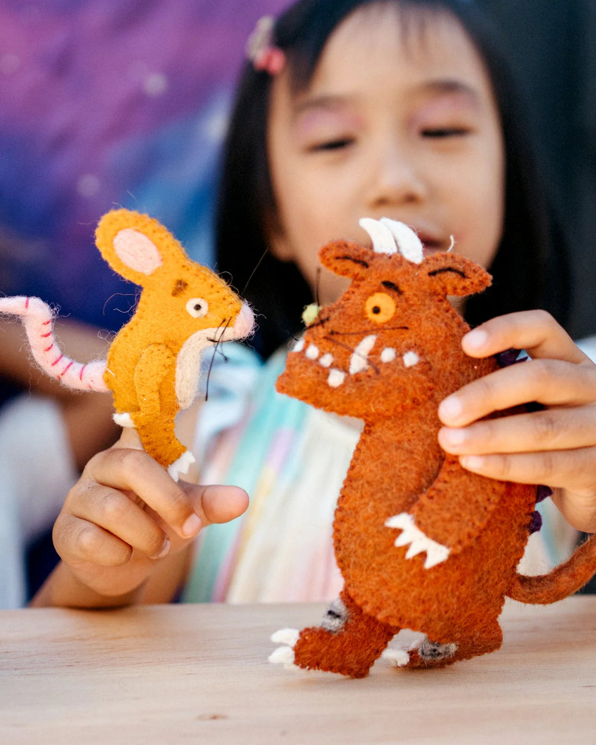 Child playing with two felt animals, a mouse and a dragon, on a wooden surface.