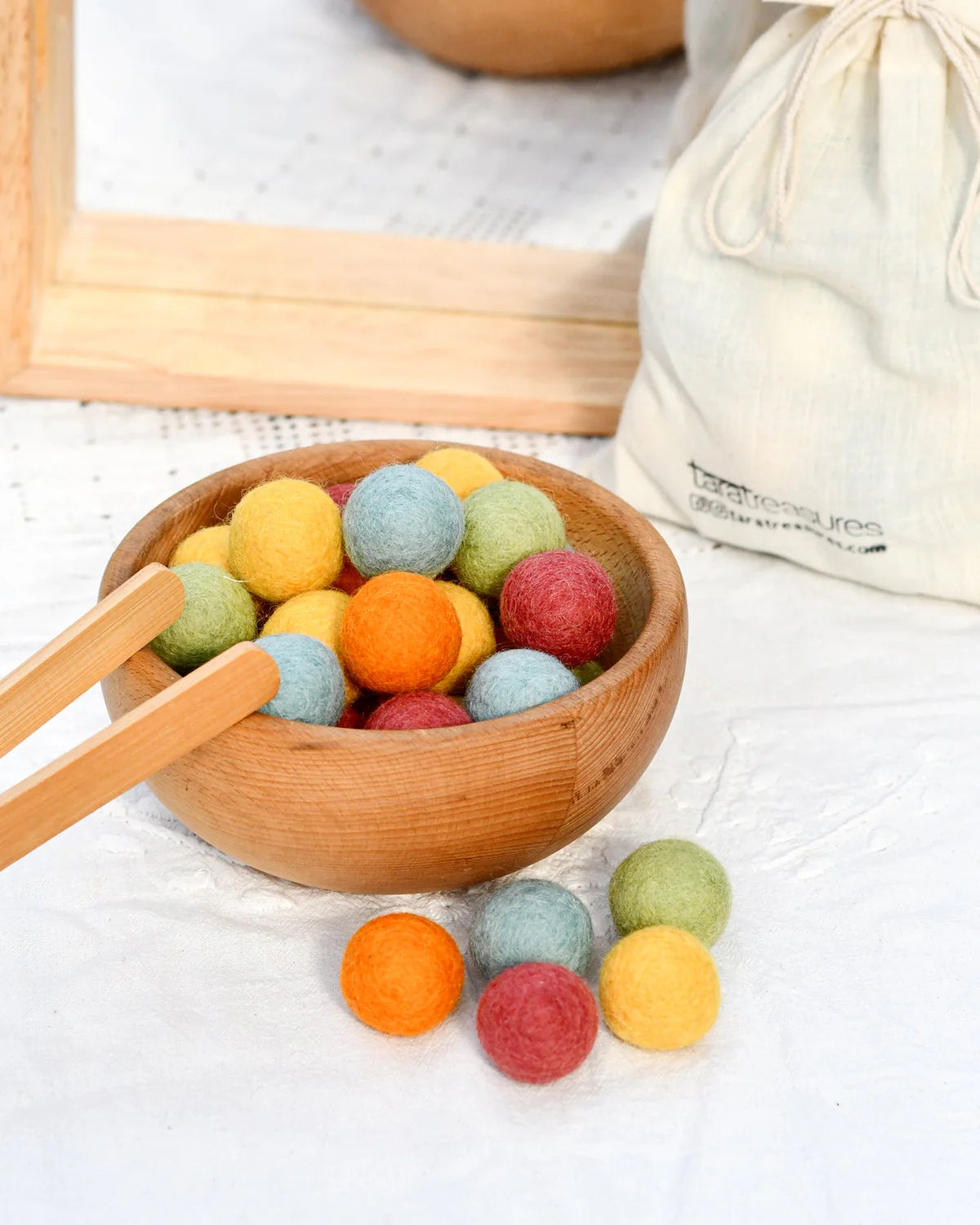 Colorful felt balls in a wooden bowl with wooden tongs on a light background