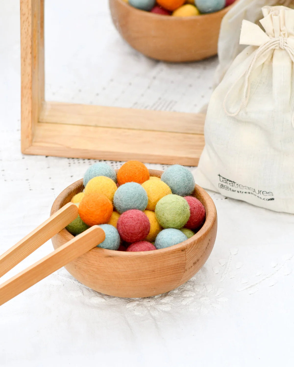 Colorful felt balls in a wooden bowl with a wooden frame and white cloth bag in the background.