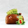 A brown felt log-shaped pouch with embroidered flowers, green felt frog characters sitting inside, and one larger green felt frog with a red number 1 on the front, set against a light gray background.