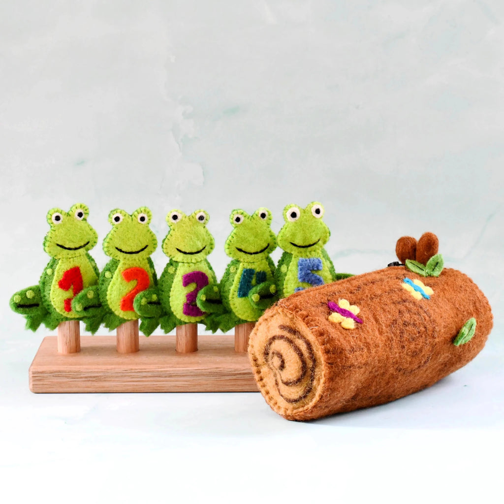 Set of green felt frogs with numbers on a wooden stand next to a brown log with colorful decorations.
