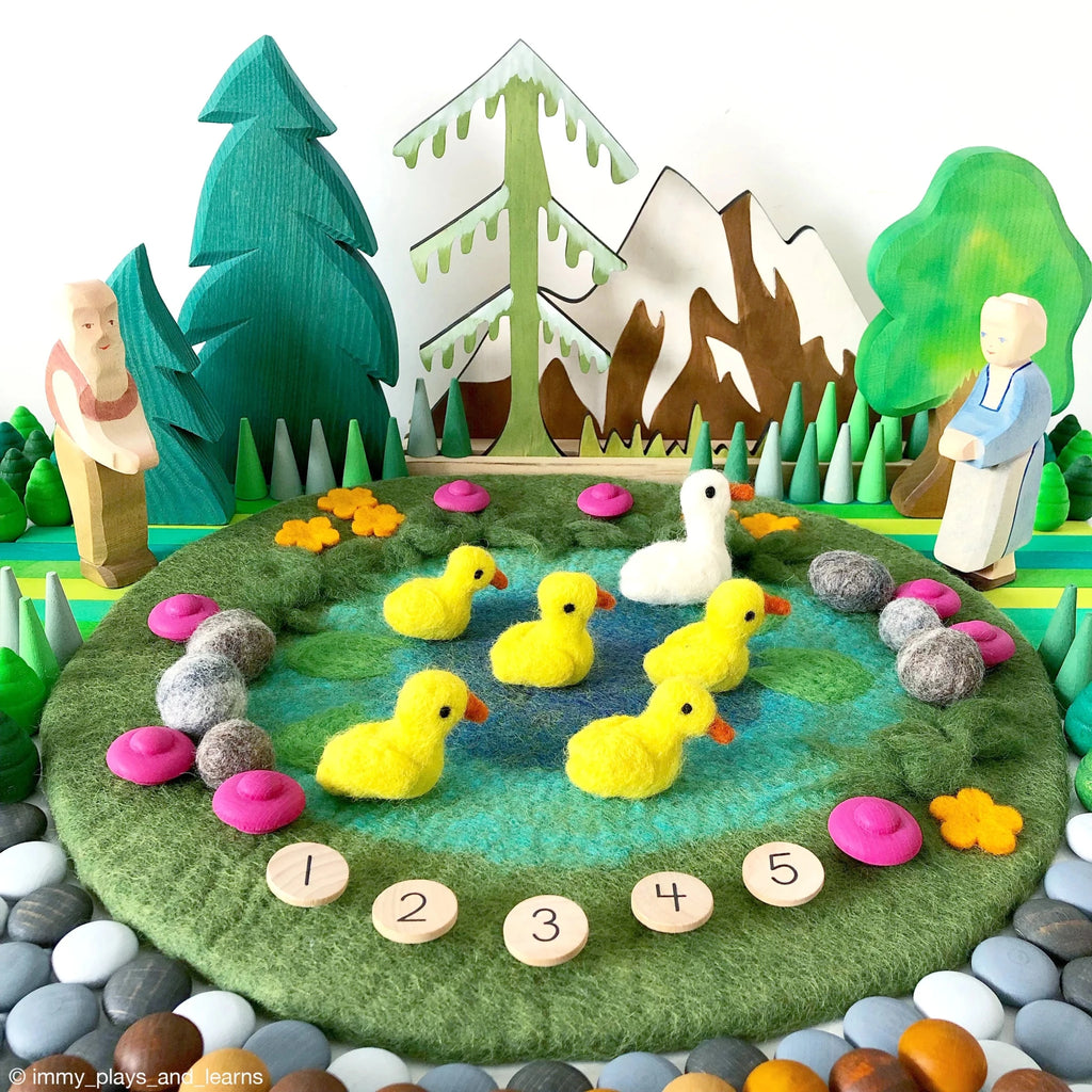 Wooden duck family and landscape with trees, flowers, and mountains on a felt surface.