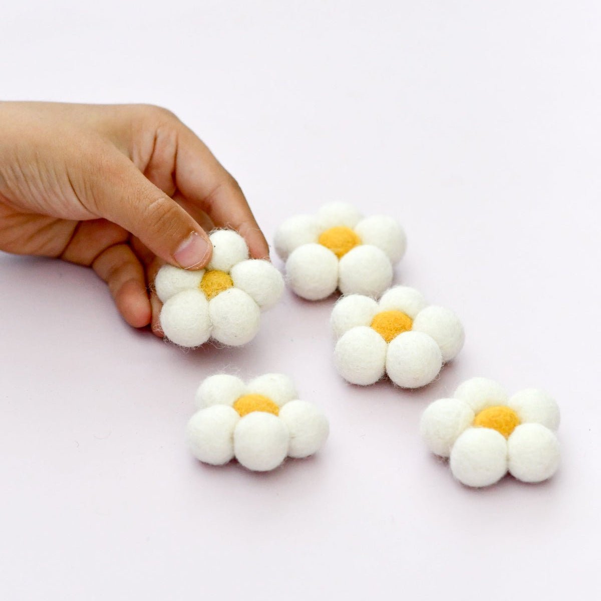 Hand holding a white flower-shaped object with a yellow center on a light pink background