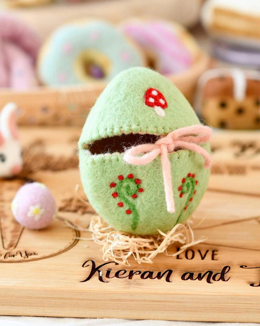 Green plush toy with a pink bow on a wooden surface, surrounded by pastel-colored items.