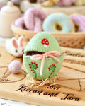 Green embroidered egg with a pink bow on a wooden board.