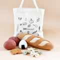 A white canvas tote bag with black food-themed illustrations and the brand name 'taratreasures' printed on it, styled with felt play food items including a baguette, potatoes, an egg, and cookies in front of it on a warm beige background.