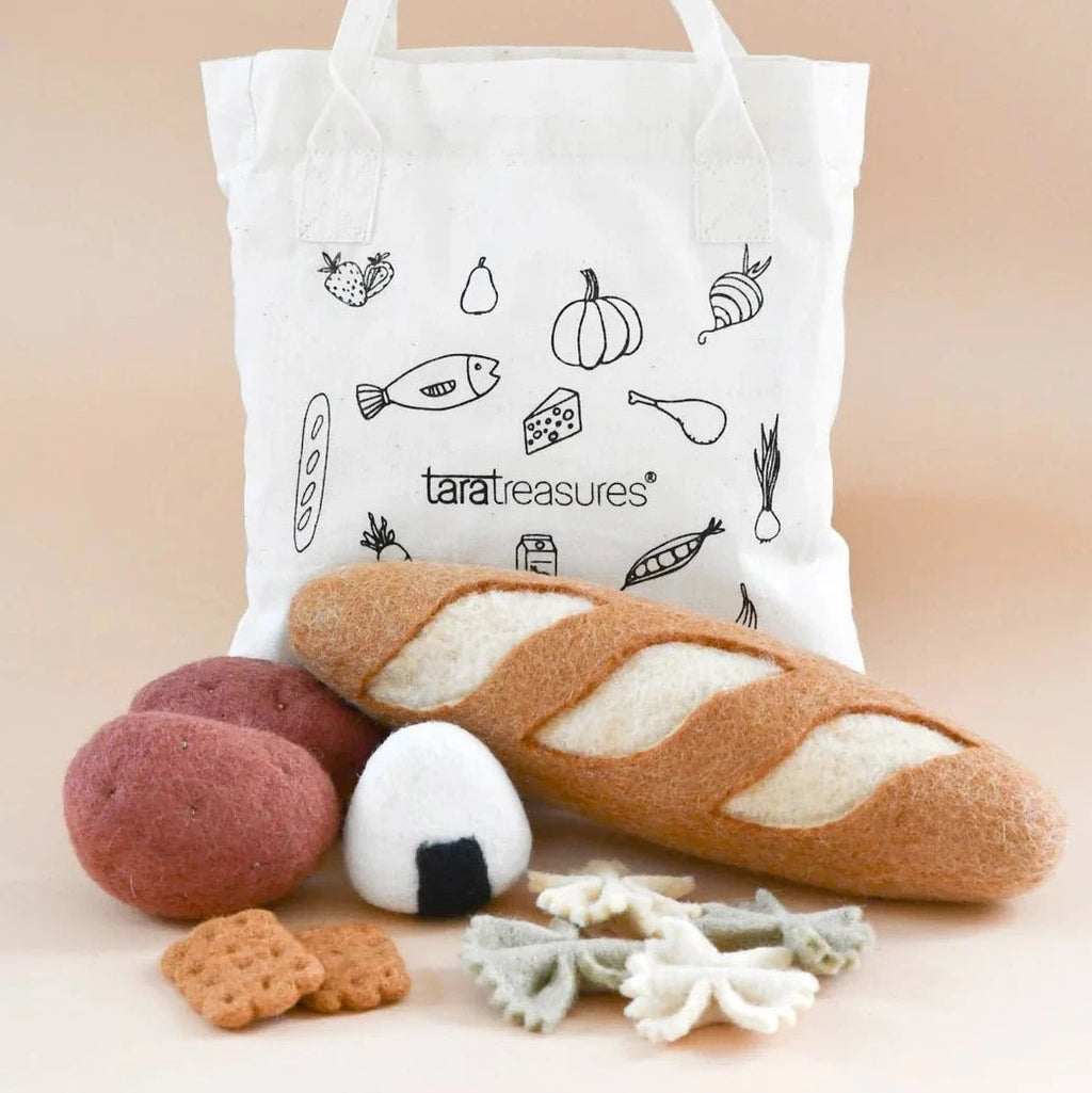 A white fabric tote bag with black line drawings of food items and the brand name 'taratreasures' printed on the front, styled with miniature felt toys resembling a baguette, an egg, breadsticks, and pasta on a warm beige background.