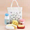 Toy food items including a bottle, cheese, and bread on a beige background with a tote bag featuring illustrations and text.