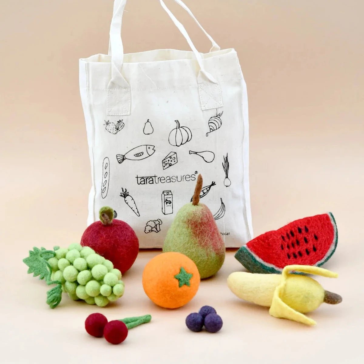 A white canvas tote bag with black line drawings of food items and the printed brand name 'tara treasures' stands behind a set of felt fruit toys including a pear, watermelon slice, banana, grapes, cherries, and an orange on a warm beige background.
