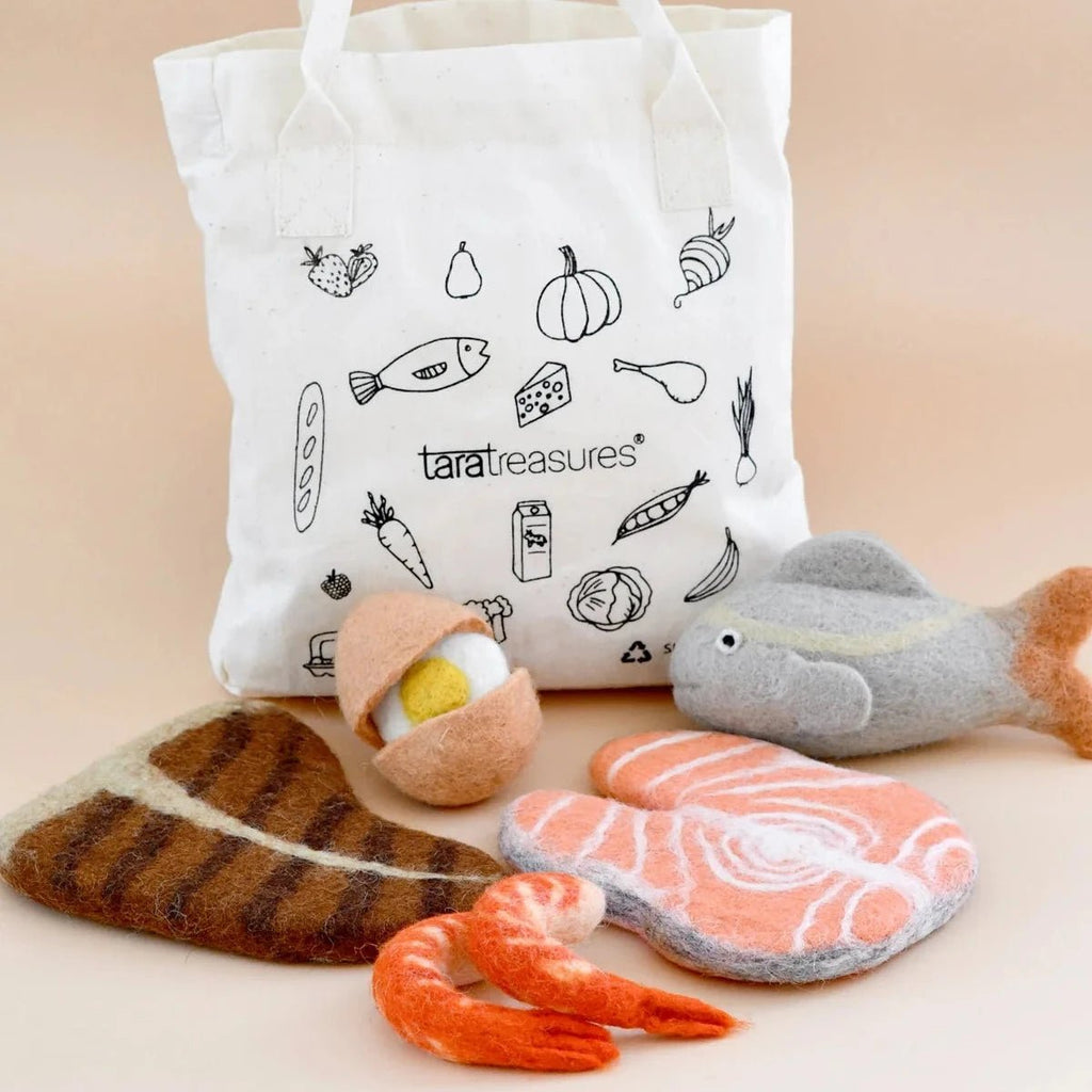 White tote bag with black illustrations and text, accompanied by plush toy food items on a beige background.