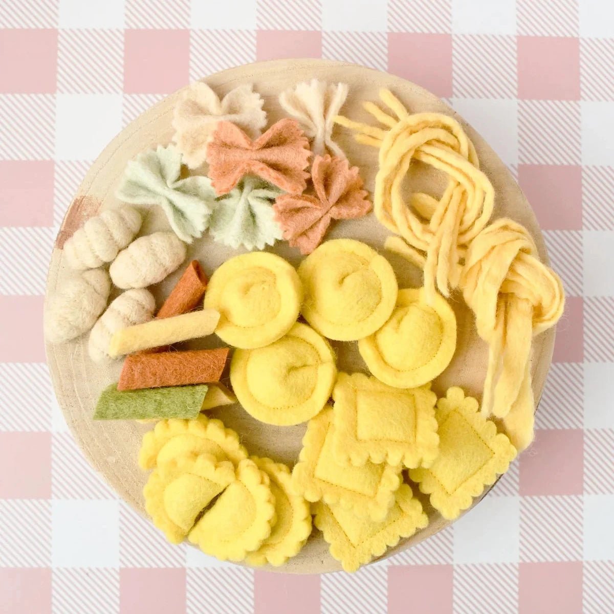A round wooden board filled with colorful felt pasta shapes, including yellow rotini, square rigatoni, bow ties, fettuccine, and other pasta designs, set against a pink and white checkered tablecloth.