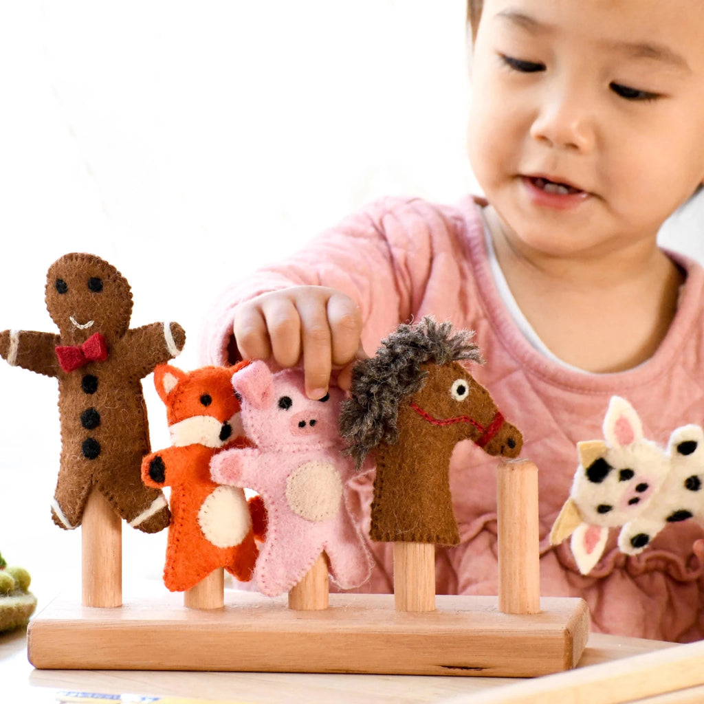 A young child wearing a pink shirt is interacting with a wooden felt puppet toy set on a light wooden table. The set includes a gingerbread man, fox, pig, horse, and cow puppets on wooden sticks mounted on a wooden base.