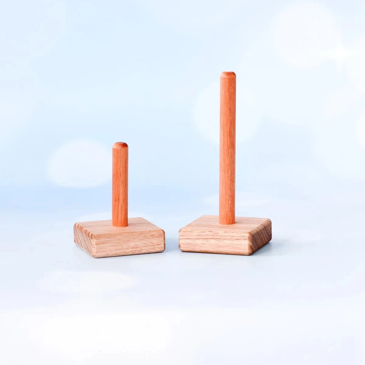 Two small wooden peg-and-base toy blocks in different heights, made of light natural wood, set against a soft light blue background with a bright bokeh effect.