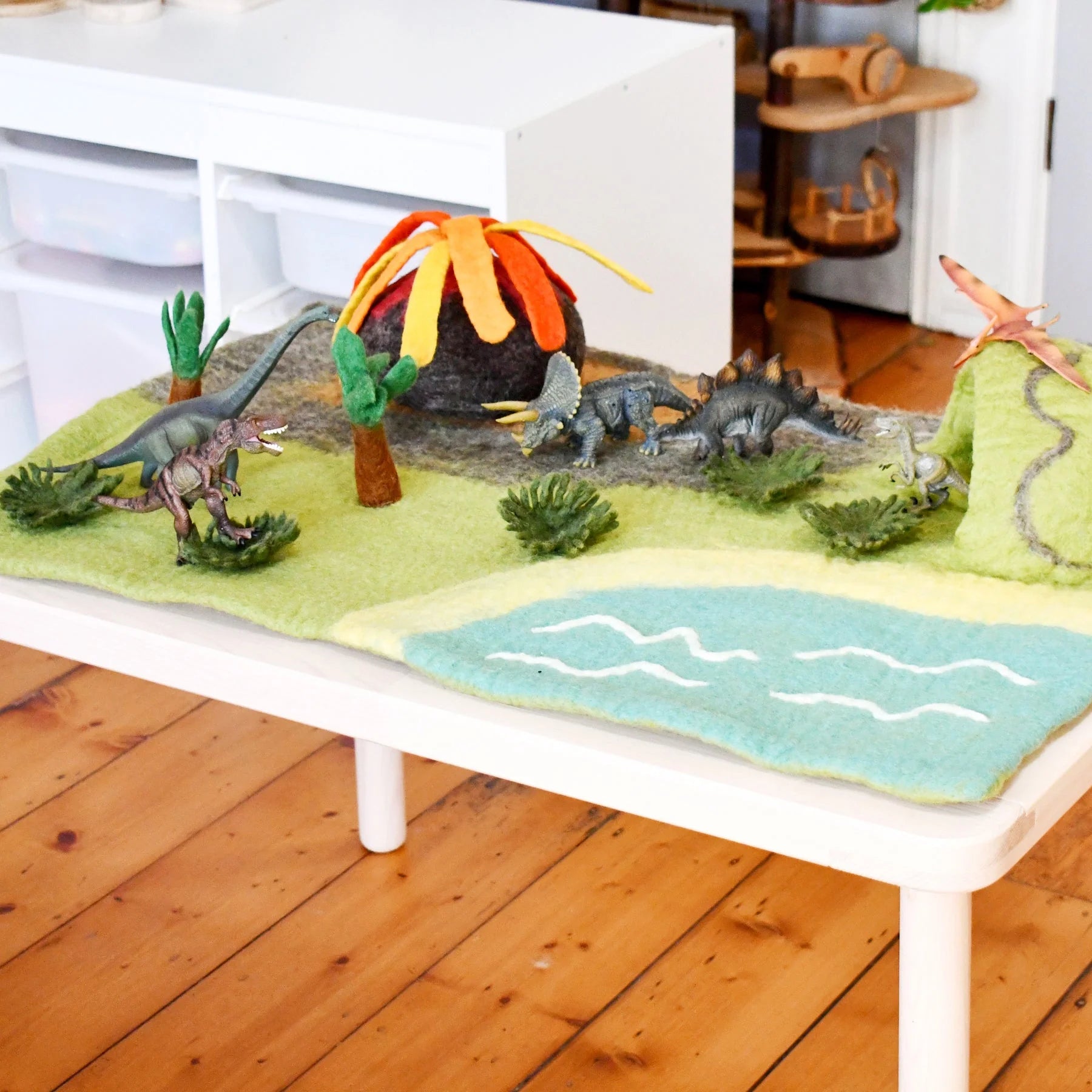 Dinosaur playset with a volcano, trees, and water on a felt surface.