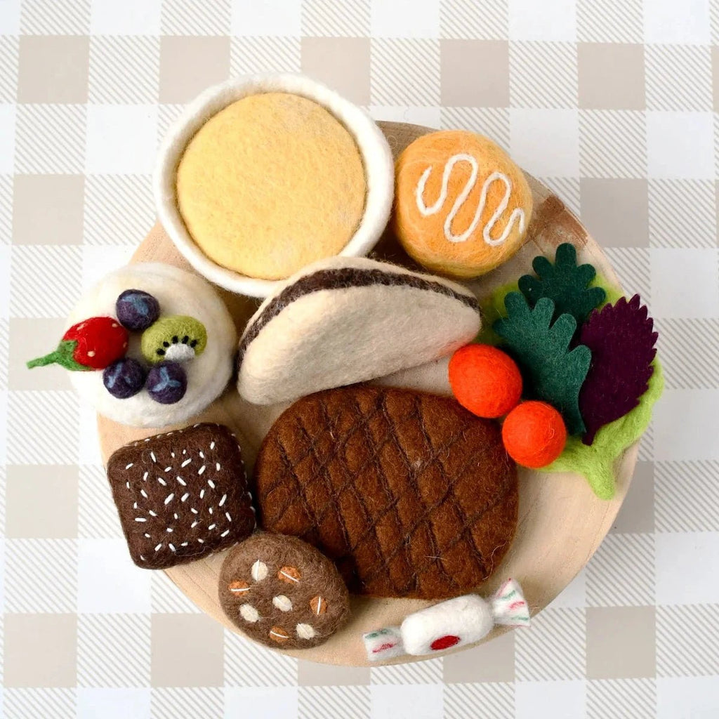 A collection of miniature felt food items arranged on a small round plate, set against a beige and white checkered background. The felt items include a hamburger, a taco, a burger, a sandwich, a cup of yellow drink, a cookie with white drizzle, a fruit salad, and various vegetables.