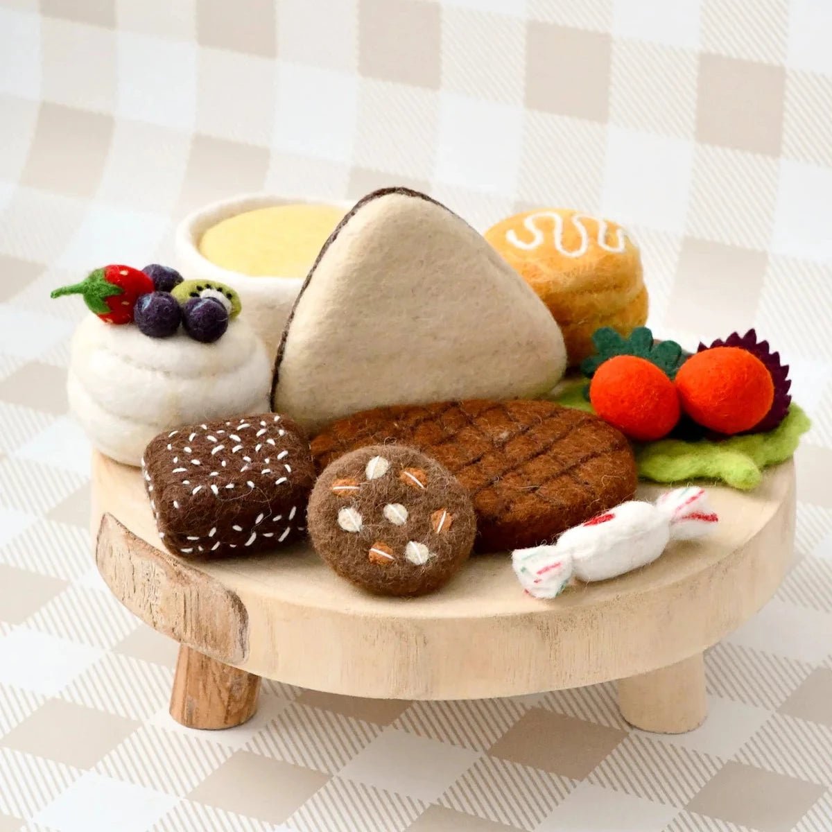 A miniature toy food set arranged on a small round wooden stand, including a triangular sandwich, cookies, fruit, a cup of yellow drink, and wrapped candy, photographed against a beige-and-white checkered backdrop.