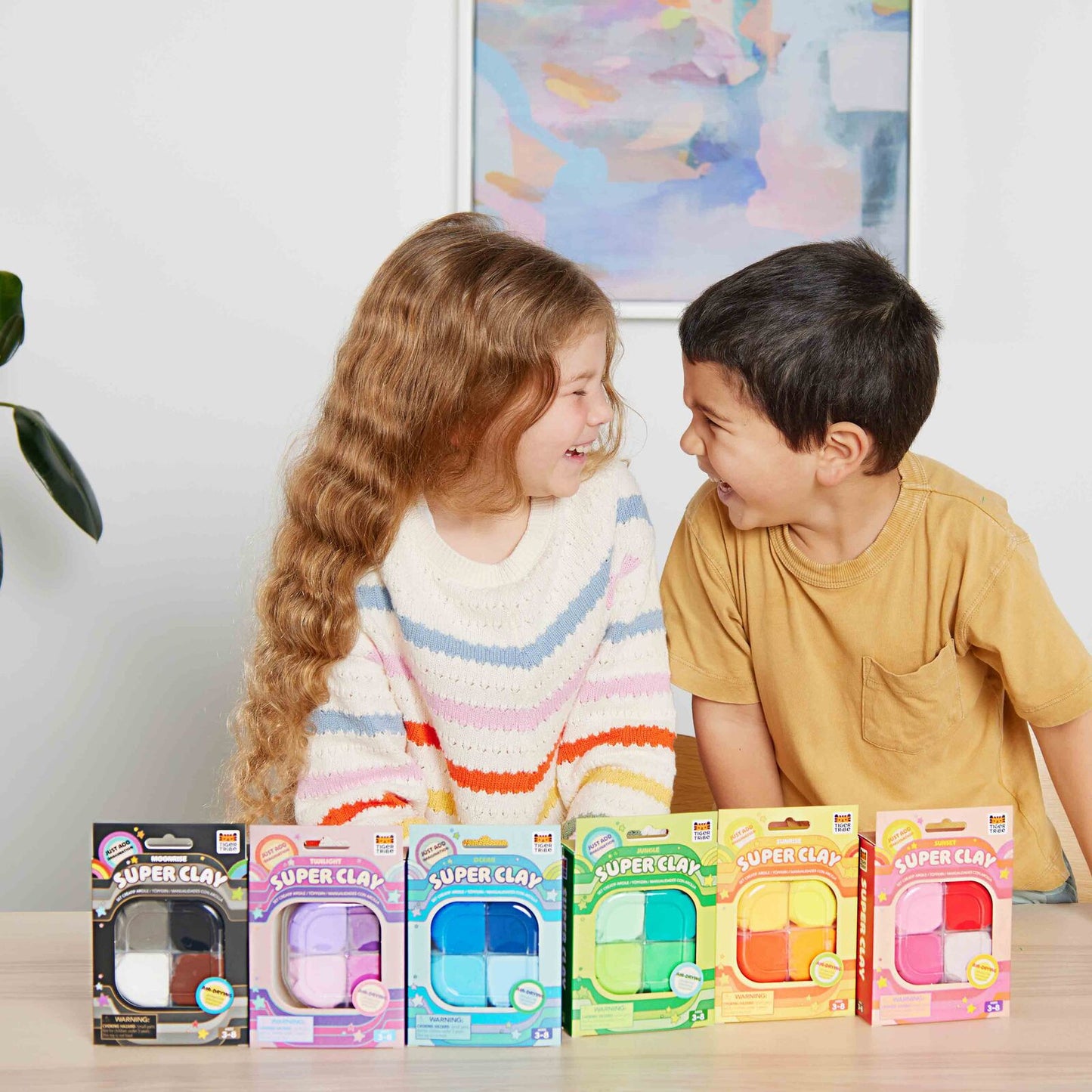 Two children sitting at a table with packages of Super Clay toys in front of them.