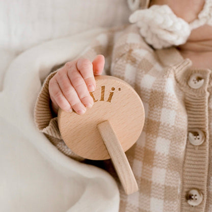 Baby hand holding a wooden rattle with 'Totli' branding, wearing a checkered outfit.