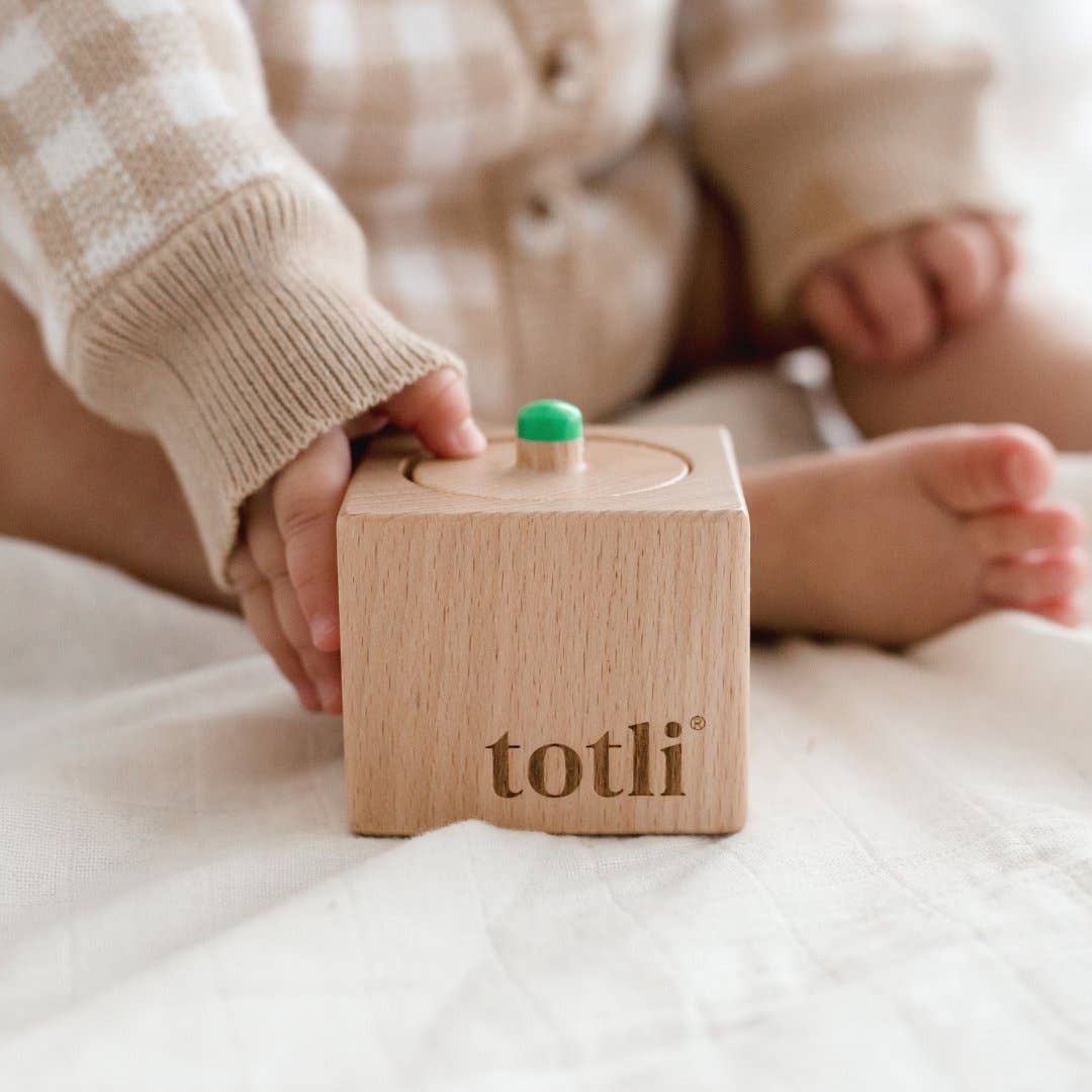 Wooden toy block with 'totli' branding held by a child's hand on a soft surface.