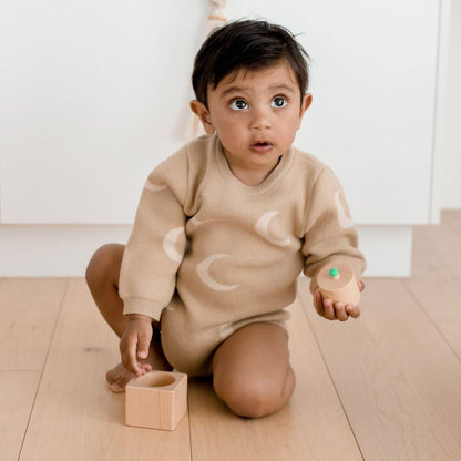 Baby in a beige outfit playing with wooden blocks on a wooden floor.