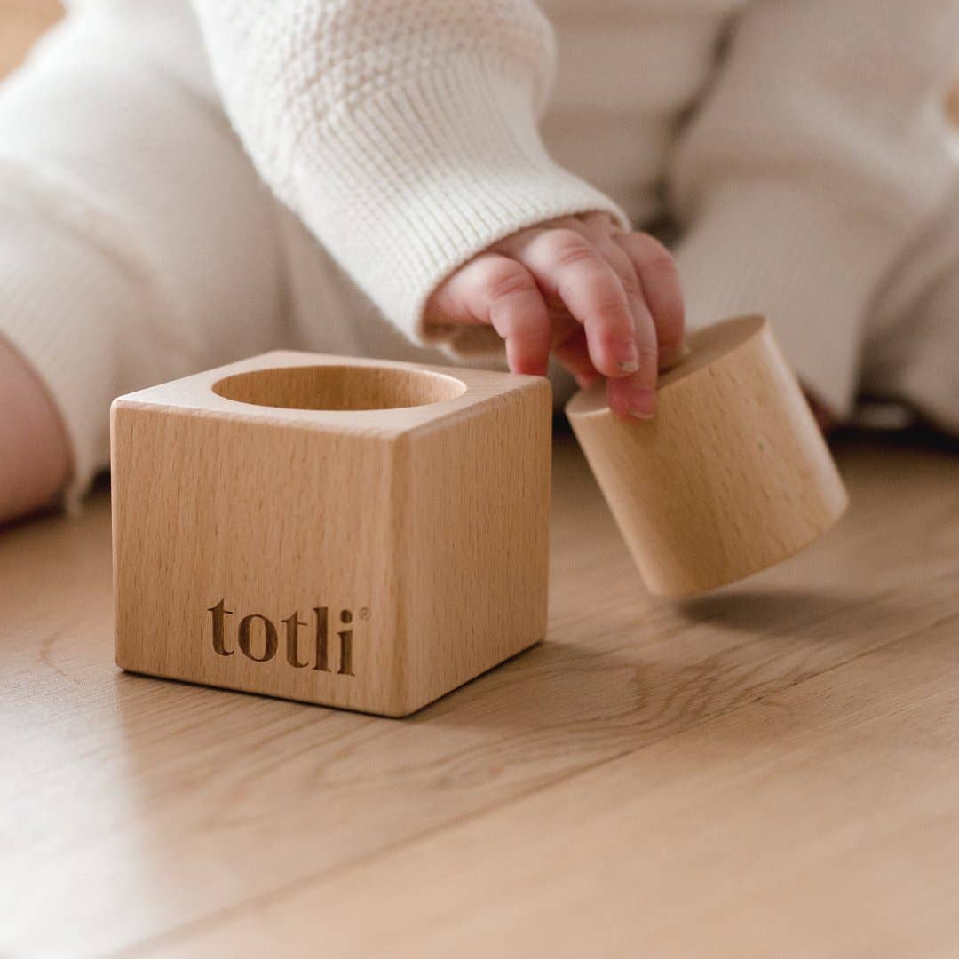 Wooden toy blocks with 'totli' branding on a wooden floor, child's hand reaching out.