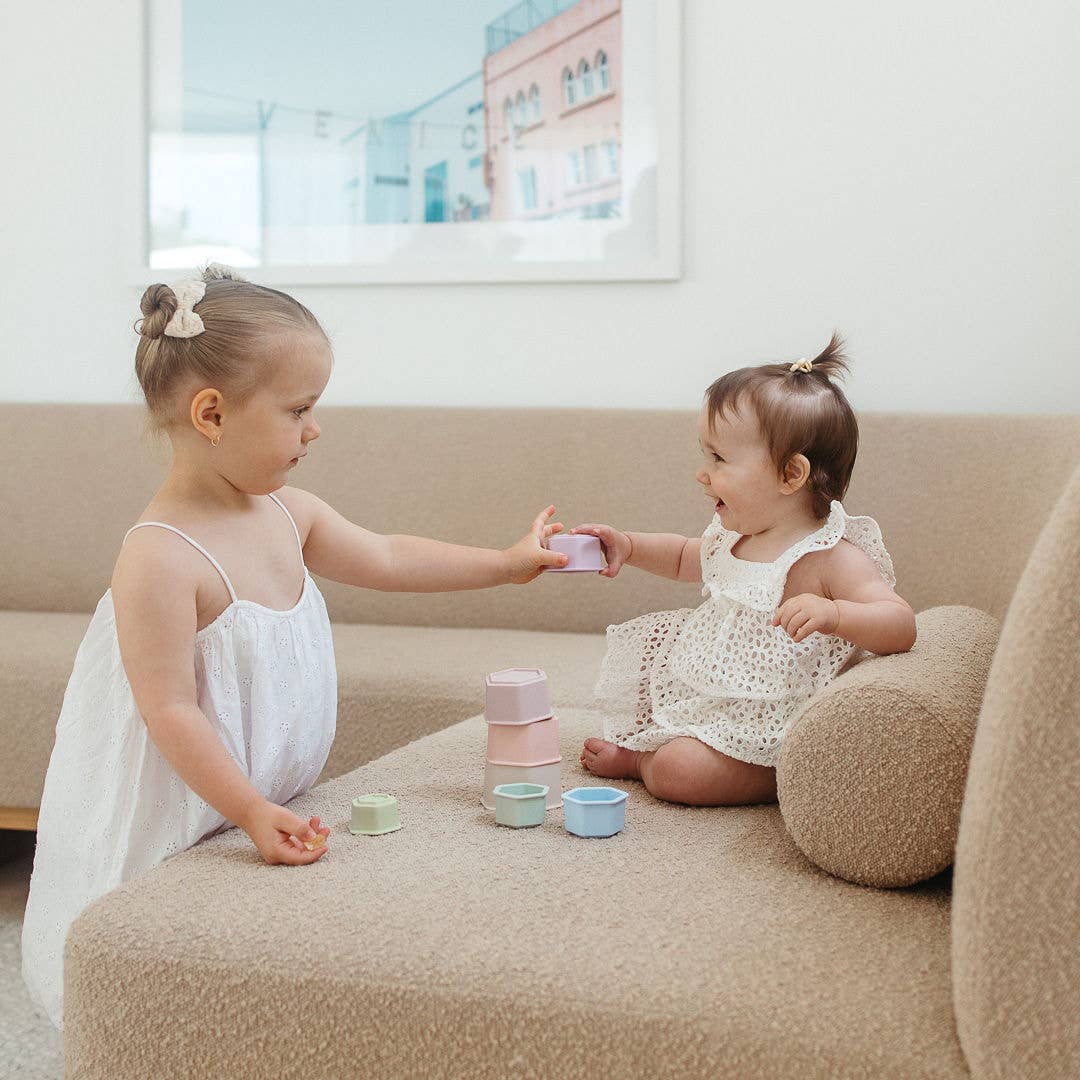 Two young girls in white dresses playing with colorful cups on a beige couch.